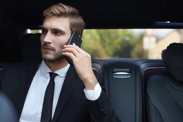 Bello uomo d'affari che parla con il telefono seduto con il computer portatile sul sedile posteriore dell'auto.