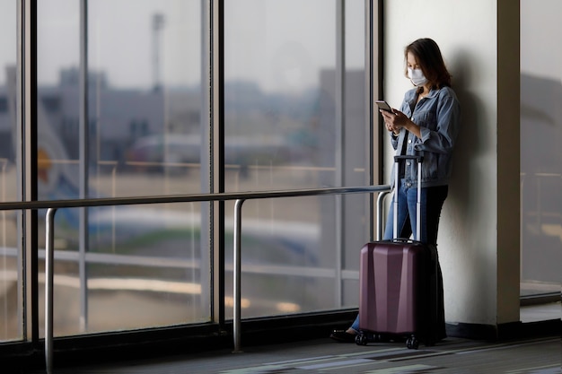 Bello viaggiatore asiatico che indossa la maschera protettiva igienica in piedi da solo e utilizza uno smartphone in aeroporto durante l'attesa di un volo. Idea per viaggiare in una nuova situazione normale.