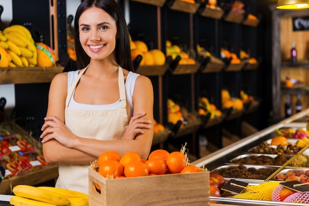 Benvenuti nel paradiso della frutta! Bella giovane donna in grembiule che tiene le braccia incrociate e sorride mentre si trova in un negozio di alimentari con una varietà di frutta sullo sfondo