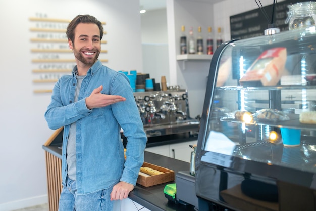 Benvenuto al caffè. Felice giovane uomo barbuto che sorride alla telecamera in piedi invitando gesticolando alla vetrina del cibo nella caffetteria