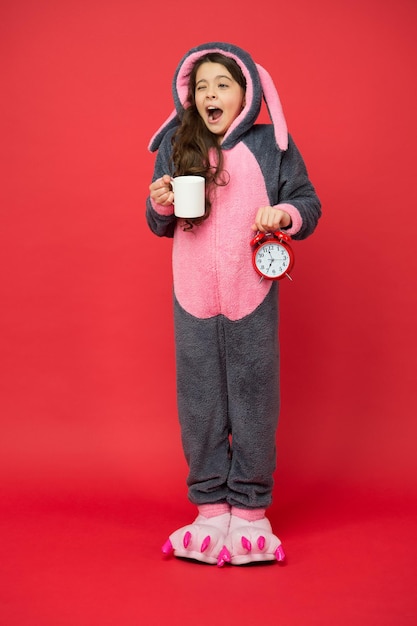 Bere energia al caffè. Routine mattutine. Adorabile sveglia a forma di coniglietto. Piccola ragazza in costume da coniglio. Kigurumi di coniglio bambino. Pigiama da coniglio per ragazza. Priorità bassa rossa del bambino del coniglietto. Animaletto. Contando il tempo.