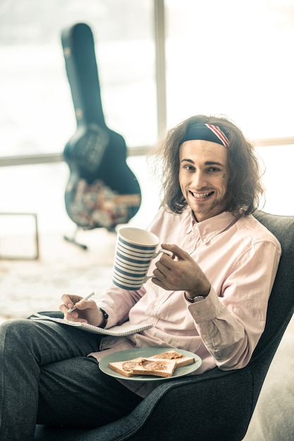 Bere il tè. Giovane contento che mostra il suo ampio sorriso mentre fa la pausa pranzo e mangia panini