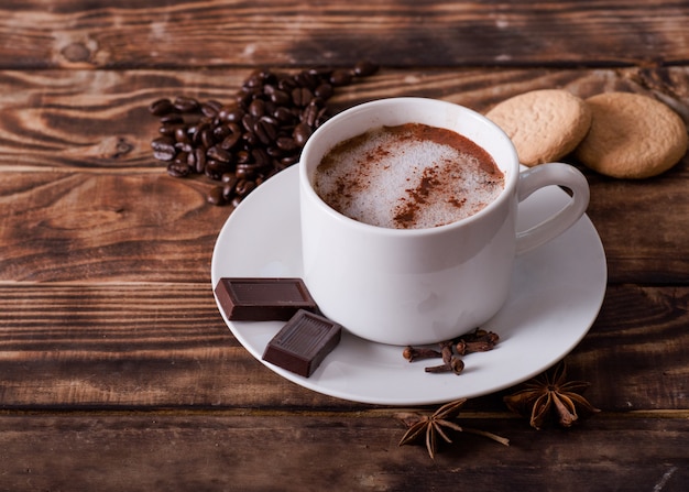 Berretto di caffè con schiuma, torte, chicchi di caffè a forma di cuore e cioccolato sul tavolo di legno