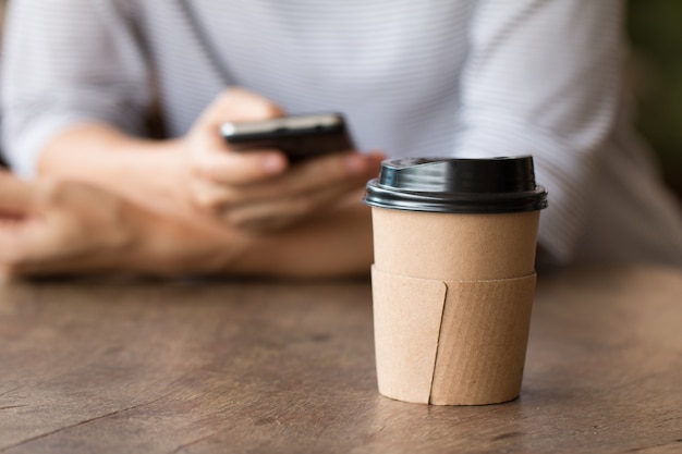 Bevanda calda della tazza di caffè di carta sullo scrittorio di legno con la donna che lavora allo Smart Phone