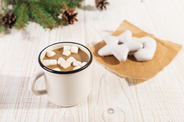 Bevanda calda di Natale con marshmallow in una tazza di ferro e biscotti di pan di zenzero, su un tavolo bianco.