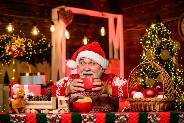 Bevanda calda. Ricetta natalizia al cacao. Ghirlanda illumina un'atmosfera accogliente. Casa accogliente. Uomo anziano Babbo Natale che beve tè. Capodanno. Nonno con la tazza. I dolcetti della vigilia di Natale. Interni accoglienti. Vacanze invernali.