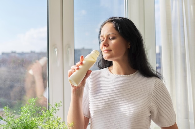 Bevanda del latte della donna dalla bottiglia che sta vicino alle bevande sane della latteria del yogurt del latte della finestra