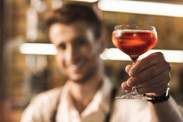 Bevanda gustosa. L'attenzione si concentra sul bicchiere con cocktail nelle mani di un piacevole giovane che lavora come barista e sorridente