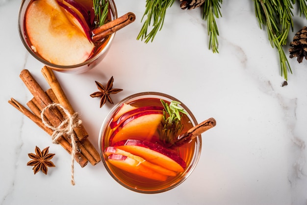 Bevande del Ringraziamento di Natale Autunno inverno cocktail grog sangria calda vin brulè - mela rosmarino anice cannella sul tavolo di marmo bianco con coni rosmarino