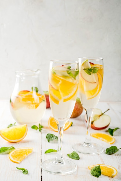 Bevande e cocktail Sangria autunnale bianca con mele, arance, menta e vino bianco. In bicchieri per champagne, in una brocca, su un tavolo di legno bianco. Copia spazio