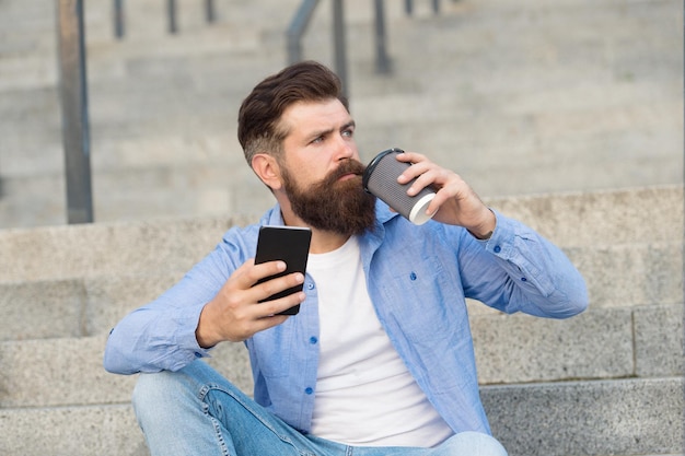 Bevilo in movimento Bere caffè brutale hipster seduto sulle scale Uomo barbuto godersi una bevanda calda all'aperto Bevanda da asporto Pausa caffè Vita moderna Comunicazione smartphone Sorseggiare accade