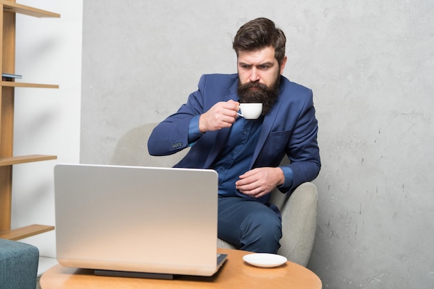 Bevo tonnellate di caffè. Uomo d'affari serio che lavora al computer portatile con una tazza di bevanda calda. Uomo barbuto del computer che si gode la sua pausa per bere qualcosa in ufficio. Duro lavoro con una bevanda gustosa.