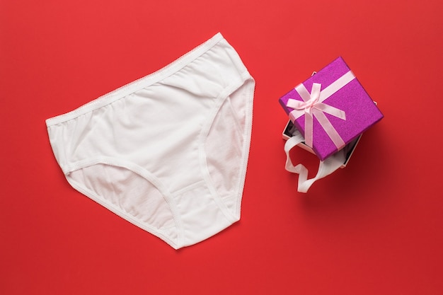 Biancheria intima da donna bianca e una confezione regalo su una superficie rossa. Disposizione piatta.