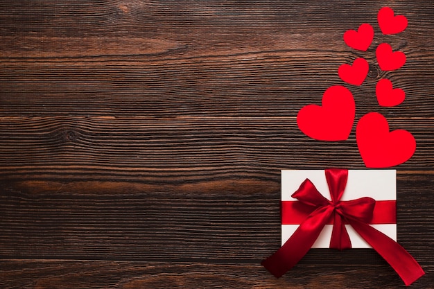 Bianco presente con un nastro rosso e cuori di carta isolati su uno sfondo di legno scuro. Vista laterale superiore di un flatlay caldo che celebra. Il giorno di San Valentino e il concetto di Natale. Copyspace.