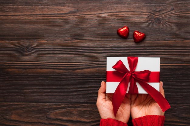 Bianco presente con un nastro rosso nelle mani della donna e due dolci al cioccolato a forma di cuore su uno sfondo di legno scuro. Concetto di giorno di San Valentino. Copyspace.