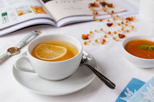 Bianco tazza di tè verde con limone, miele e menta su un lenzuolo bianco con una rivista illustrata, collana di ambra, orologio e bustine di tè. Concetto sano.