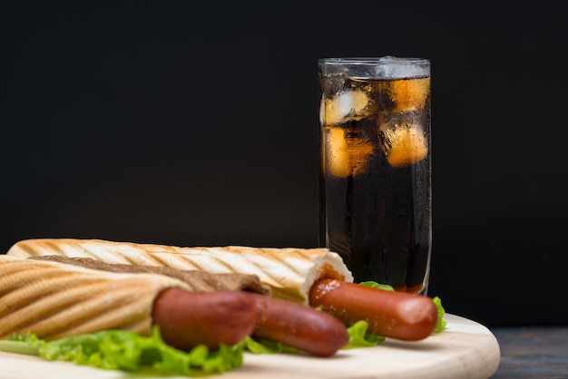 Bicchiere alto di soda con ghiaccio accanto al piatto di hot dog avvolti nel pane sopra grandi foglie di lattuga