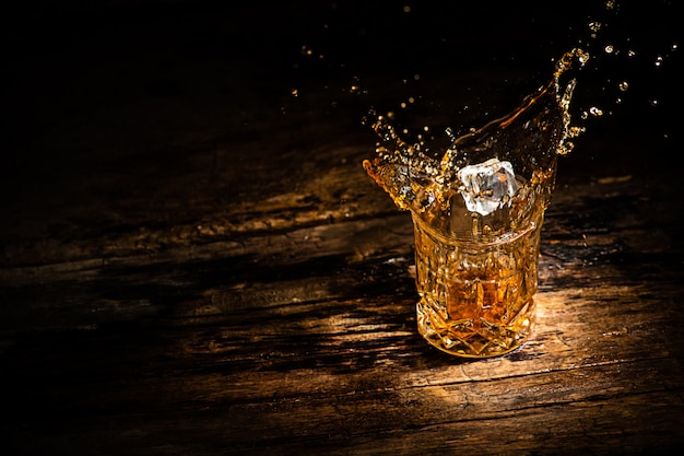 Bicchiere con whisky o cognac o un altro tipo di alcol con schizzi dal cubetto di ghiaccio su sfondo di legno