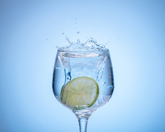 Bicchiere d'acqua con limone caduto e spruzzata su sfondo azzurro sfumato.