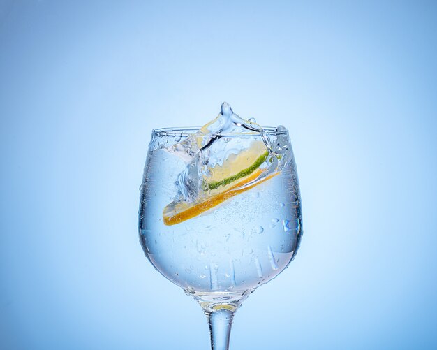 Bicchiere d'acqua con limone e palline di ghiaccio colorate su sfondo azzurro sfumato.