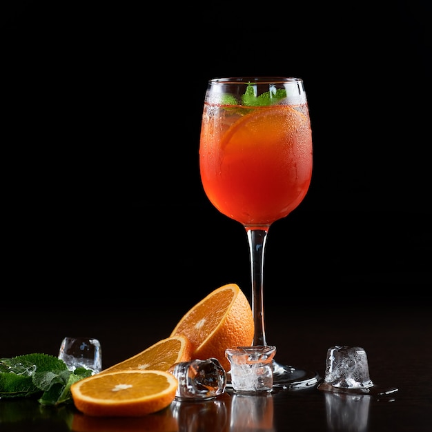 Bicchiere da cocktail in cristallo alto con bevanda succosa rinfrescante estiva fredda, fette di arancia, foglie di menta verde fresca e cubetti di ghiaccio trasparenti su sfondo nero profondo. Proporzioni immagine 1:1