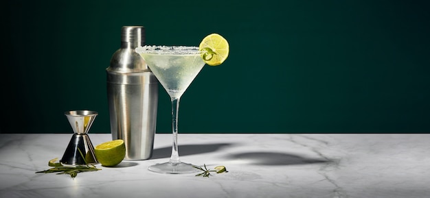 Bicchiere da Martini con cocktail o mocktail e shaker con cuneo di lime e jigger su sfondo scuro