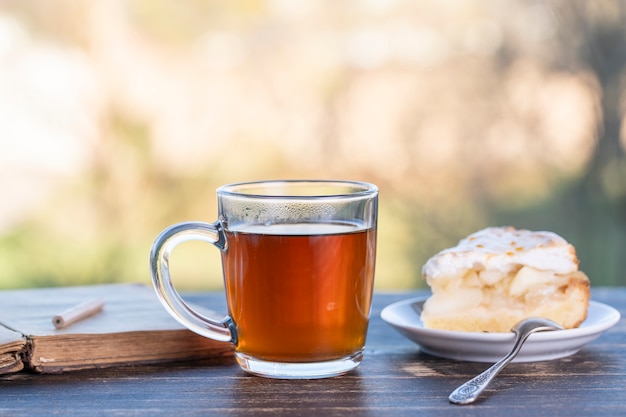 Bicchiere da tè con torta dolce e libro sul tavolo di legno al mattino, all'aperto, primo piano