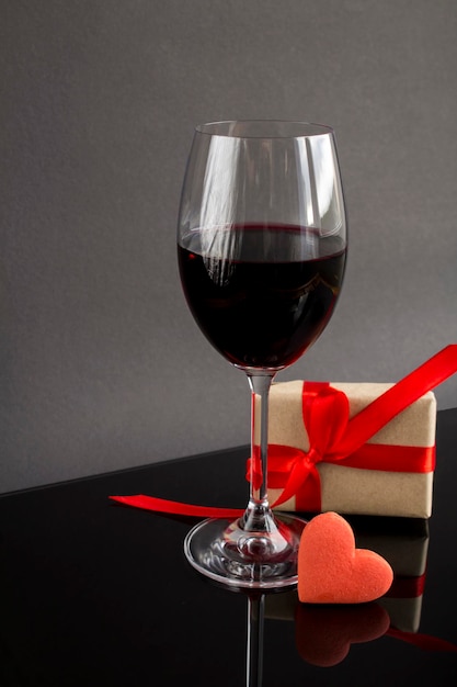 Bicchiere da vino con vino rosso, confezione regalo e biscotto a forma di cuore rosso su sfondo scuro. Avvicinamento.