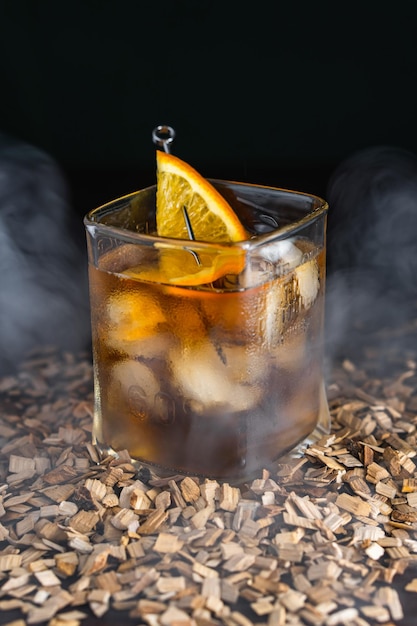 Bicchiere da whisky con cubetti di ghiaccio e fetta d'arancia su spiedino da cocktail Alcool sparato su segatura in fumo