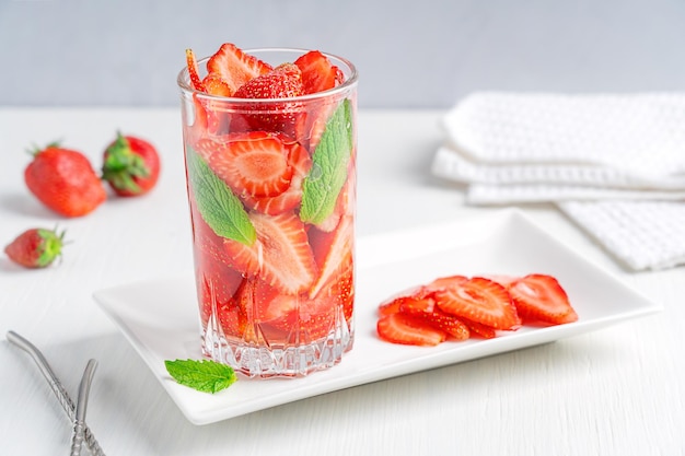 Bicchiere di acqua infusa fredda rinfrescante fatta di fragole e foglie di menta servita su un tavolo bianco