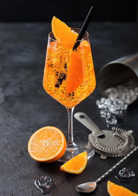 Bicchiere di aperol spritz cocktail estivo con fette d'arancia e colino con jigger e shaker su sfondo nero con cubetti di ghiaccio