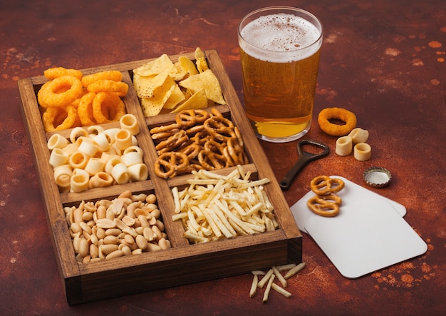 Bicchiere di birra chiara artigianale e apriscatole con scatola di snack sul tavolo da cucina marrone. Pretzel, bastoncini di patate salate, arachidi, anelli di cipolla con nachos in scatola vintage con apriscatole e tappetini per birra.