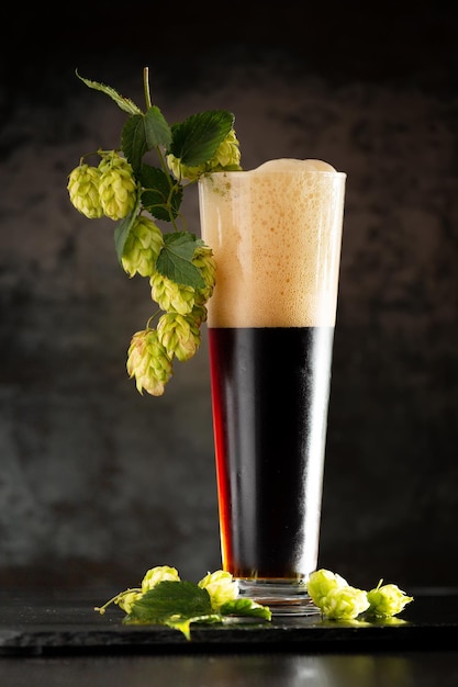 Bicchiere di birra scura con luppolo intorno su uno sfondo scuro