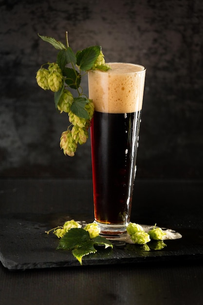 Bicchiere di birra scura con luppolo intorno su uno sfondo scuro