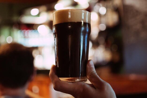 Bicchiere di birra stout scura con schiuma e bollicine in mano a un uomo nel pub dietro al bar