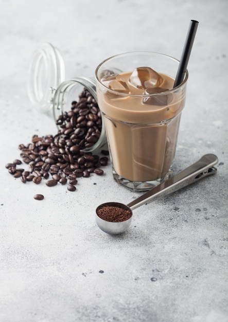 Bicchiere di caffè freddo con latte con barattolo di chicchi di caffè e cucchiaio d'argento su sfondo chiaro del tavolo.