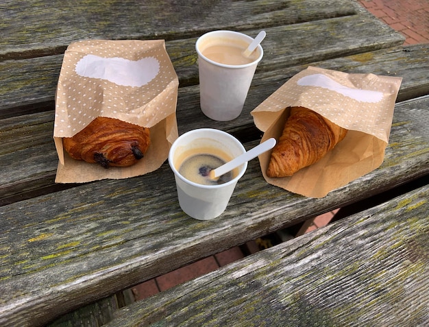 Bicchiere di carta con caffè gustoso Croissant francese pasto mattutino da asporto vecchio tavolo in legno nella vista dall'alto del parco