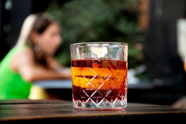 bicchiere di cocktail negroni rosso con fette d'arancia e blocco di ghiaccio trasparente sullo sfondo della tavola in legno tavolo donna sfocata