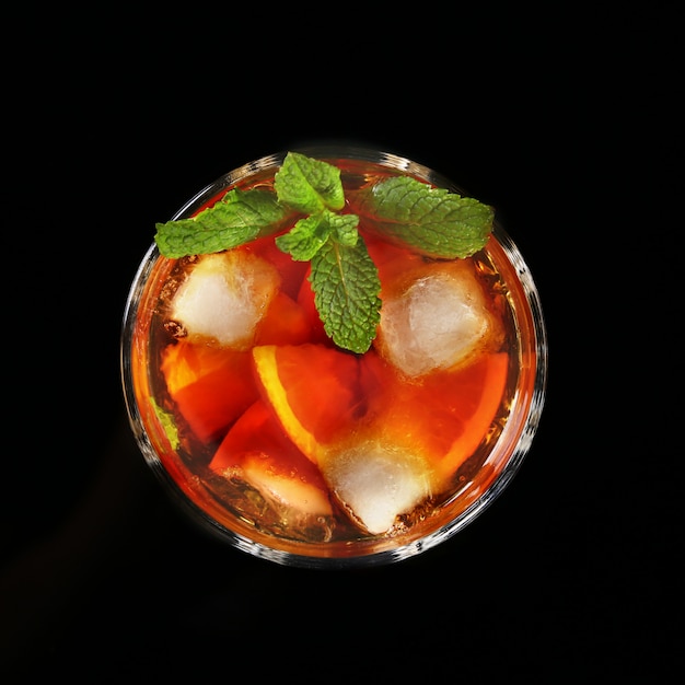 Bicchiere di Rum scuro Cocktail con lime, arancio, cubetti di ghiaccio e foglie di menta su sfondo nero a specchio.