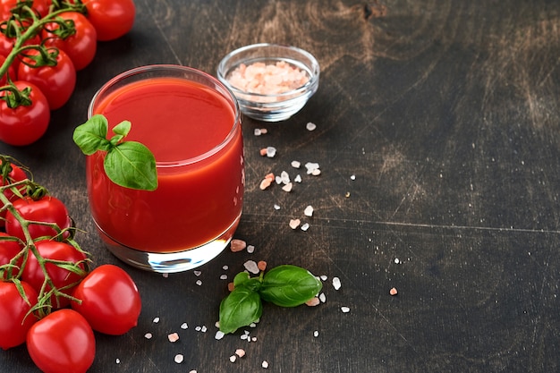 Bicchiere di succo di pomodoro fresco, sale, basilico e pomodori su supporto in legno su fondo di legno vecchio. Con copia spazio.