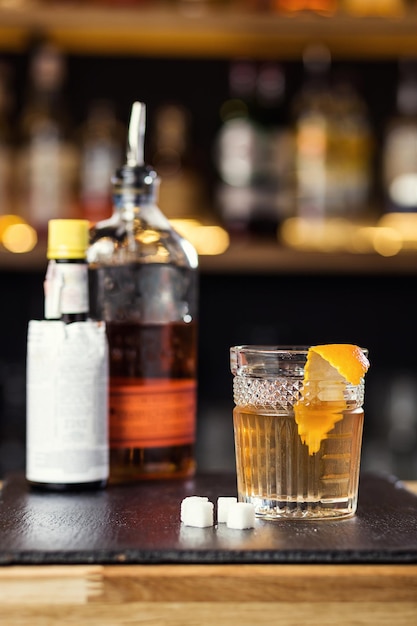 Bicchiere di whisky cognac o brandy con limone e cubetti di ghiaccio in piedi sul bancone del bar con una bottiglia sullo sfondo