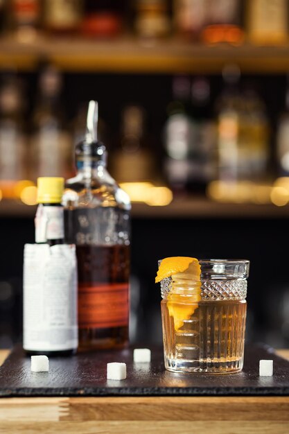Bicchiere di whisky cognac o brandy con limone e cubetti di ghiaccio in piedi sul bancone del bar con una bottiglia sullo sfondo