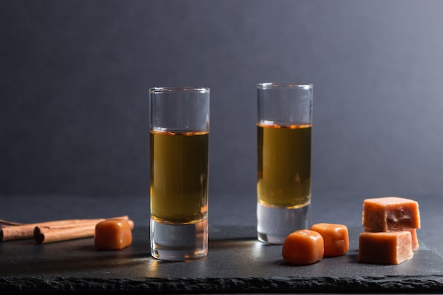Bicchiere di whisky con caramelle al caramello su una tavola di pietra nera ardesia sulla superficie nera. Vista laterale.