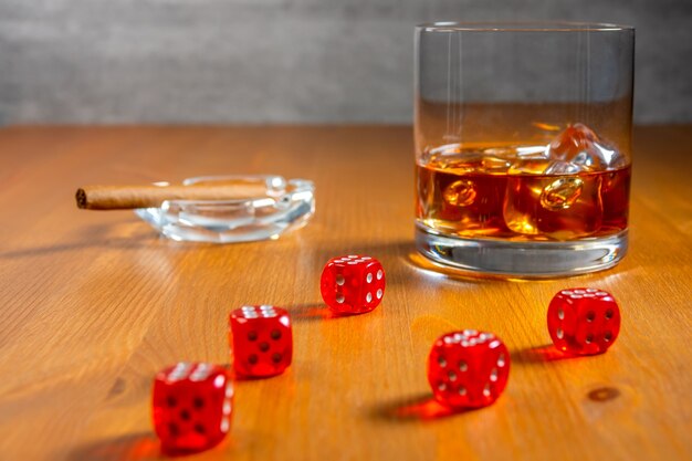 Bicchiere di whisky con cubetti di ghiaccio e posacenere con sigaro. Dadi rossi su un tavolo di legno