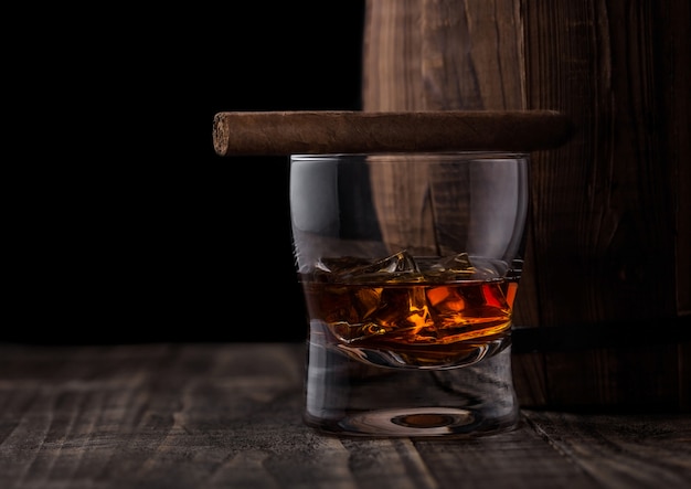 Bicchiere di whisky con cubetti di ghiaccio e sigaro accanto alla botte di legno