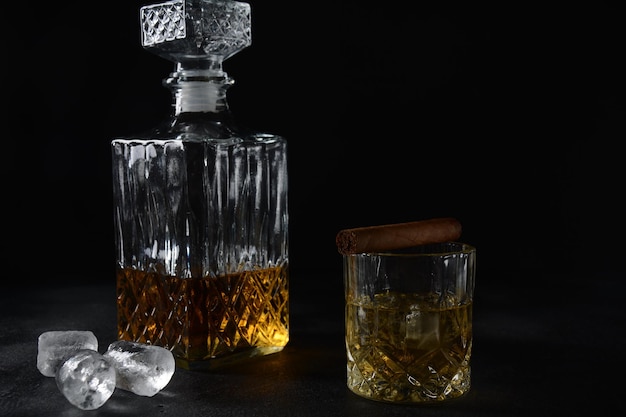 Bicchiere di whisky con cubetti di ghiaccio e un decanter quadrato e sigaro su sfondo nero.