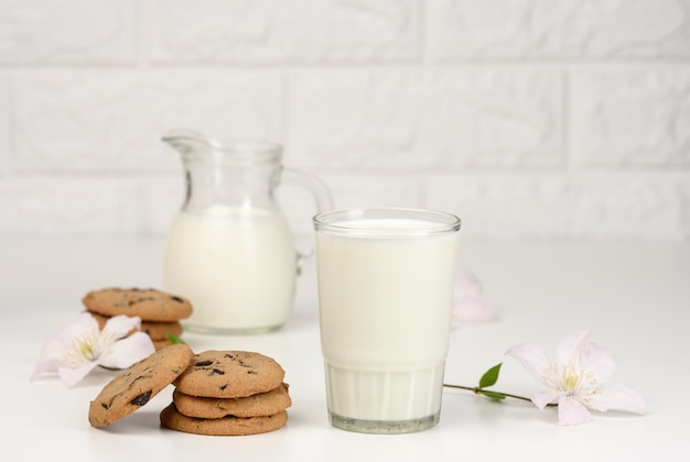 Bicchiere pieno di latte e pila di biscotti rotondi con cioccolato sul tavolo bianco