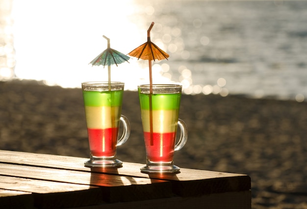 Bicchieri con cocktail multicolori su un tavolo di legno sotto il sole, la spiaggia e il mare