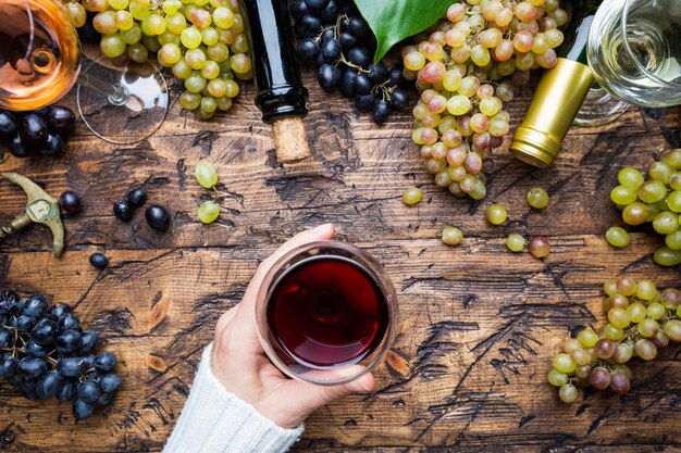 Bicchieri con vino bianco, rosso e rosa e uva matura su fondo di legno, vista dall'alto. mano di donna con bicchiere di vino