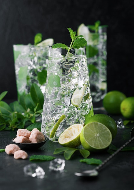 bicchieri di cocktail Mojito con cubetti di ghiaccio menta e lime su tavola nera con cucchiaio e lime fresche con zucchero di canna la migliore bevanda da festa